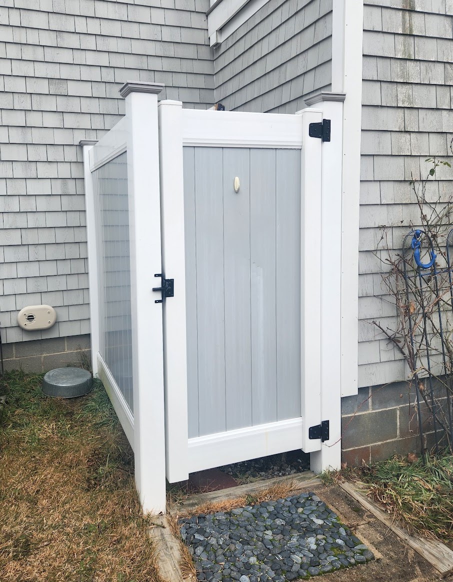 White vinyl outdoor shower enclosure with privacy panels and a gate, positioned on a deck attached to a Cape Cod style house with gray cedar shingles. The enclosure stands on a stone path surrounded by grass, with a hose reel and some plants nearby. The setting feels clean, private, and welcoming. No visible text.